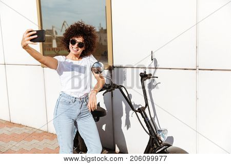 Smiling pretty girl in sunglasses taking a selfie while leaning on a motorbike outdoors