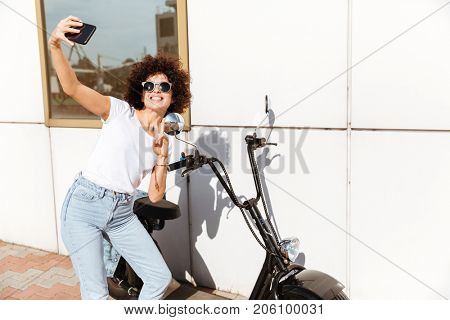 Smiling young girl in sunglasses taking a selfie and showing victory gesture while leaning on a motorbike outdoors