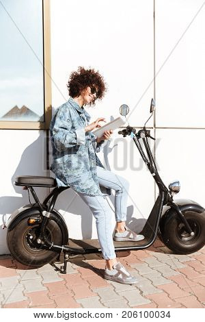 Smiling pretty woman in sunglasses using tablet computer while sitting on a motorbike outdoors