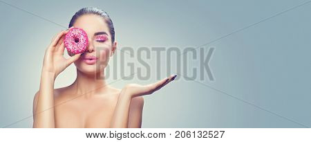Beauty fashion model girl holding sweet pink donut and pointing hand, showing products. Advertising. Vogue styled woman with pink doughnut and sugar sprinkles makeup. Fashion art make up. Diet
