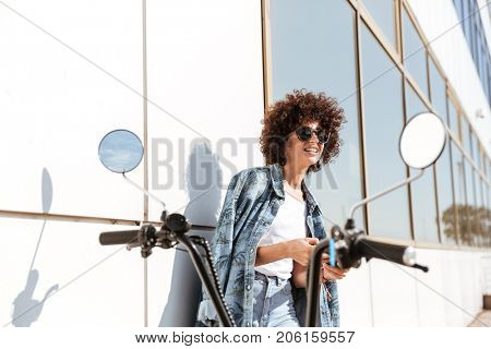 Stylish pretty woman in sunglasses using mobile phone while standing against the wall outdoors with a motorbike