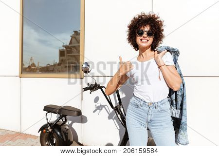 Happy cheerful woman in sunglasses posing while standing against a wall outdoors with a motorbike and showing thumbs up gesture