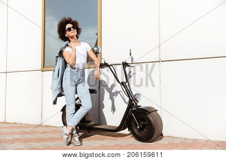 Young attractive woman in sunglasses posing while standing against a wall outdoors with a motorbike
