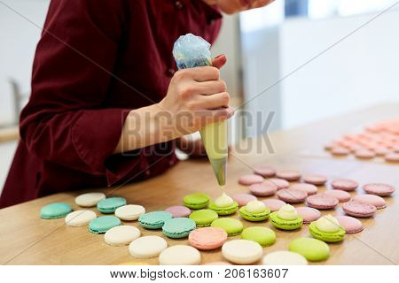 cooking, food and baking concept - chef with confectionery bag squeezing cream filling to macarons shells at pastry shop
