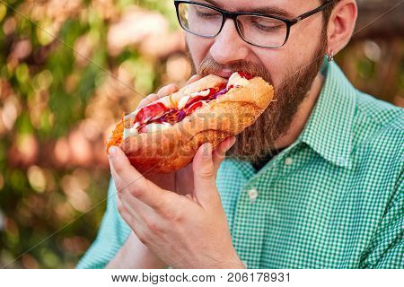 Handsome man with beard eating juicy hotdog with ketchup and majo on the street. Close-up of fast food. Food concept.