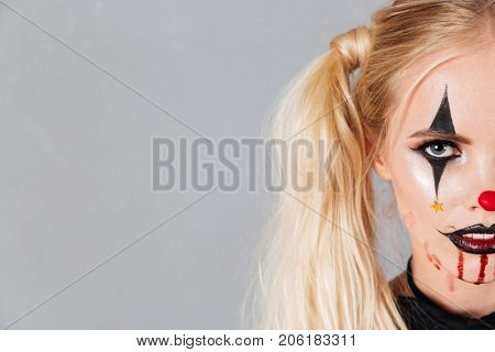 Half face portrait of a young blond woman in halloween clown make-up and blood streaks on her face looking at camera isolated over gray background