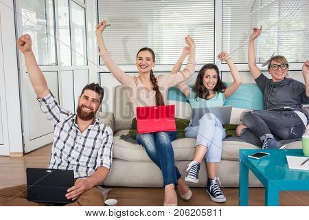 Group portrait of four happy and successful remote workers looking at camera with raised arms while sharing the facilities of a modern co-working space