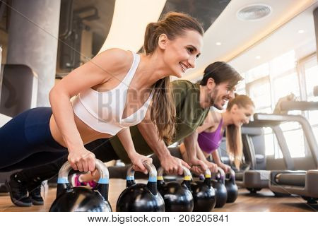 Side view of three young people smiling while doing the kettlebell plank challenge for strong core during group workout at the gym