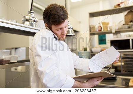 cooking, profession and people concept - male chef cook with clipboard calling on smartphone at restaurant kitchen
