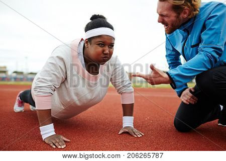 Dedicated sport trainer encouraging overweight young woman try harder while practicing push-ups