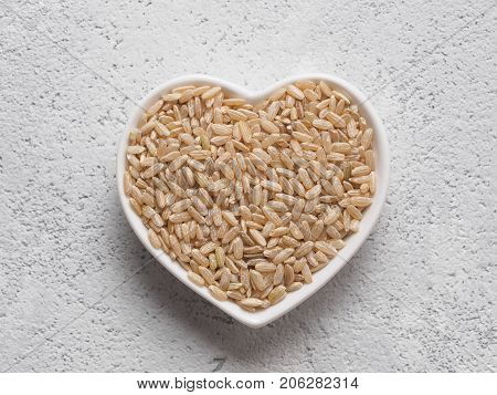 Brown rice in heart-shaped bowl on gray cement background. Copy space. Top view or flat lay.