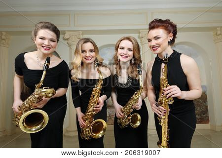 Four beautiful women in black dresses with saxophones smiles in studio with pilasters