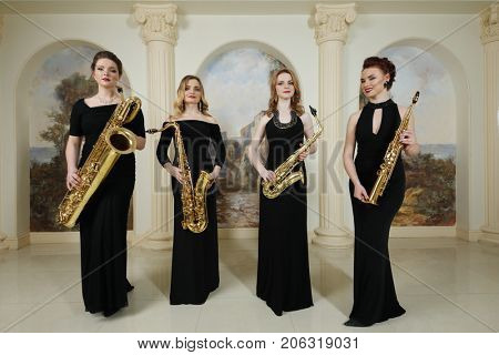 Four women in black dresses stand with saxophones in studio with pilasters