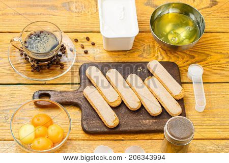 Photo of ingredients for dessert, savoyardi cookies on cutting board, coffee, cream cheese, yolks, proteins