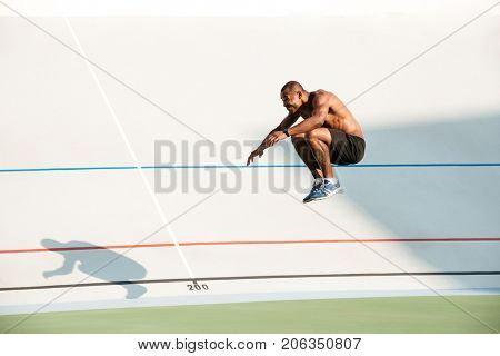Full length portrait of a half naked fit african sportsman doing a high jump at the track field outdoors