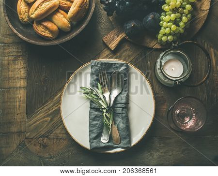 Fall holiday table decoration setting. Plate with linen napkin, fork and spoon, candle, fresh fruits on serving board, bread buns, olive tree branch over wooden background, top view, copy space