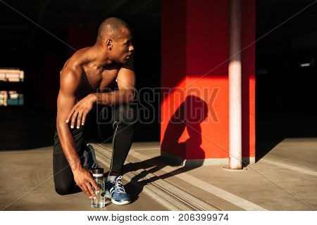 Portrait of a fit muscular half naked african sportsman sitting on his knee and resting indoors