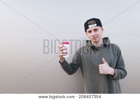 A handsome young man with a cup of coffee in his hands shows a thumbs up on a light background.