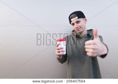 A stylish young man with a cup of coffee in his hands shows a thumbs up on a light background. The student likes coffee.