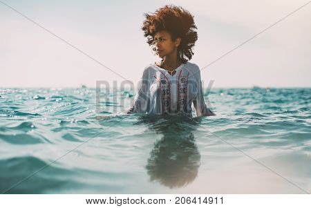Attractive young black curly girl in traditional African chemise is standing up to the waist in ocean water and looking aside strong reflection in the bottom ripples on water surface