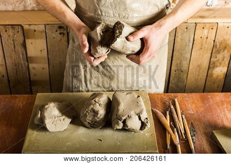 pottery, clay, ceramics art concept - top view on hands of young master holding the large pieces of fireclay, male dressed in an apron, a ceramist with raw materials on table with sculpting tools set