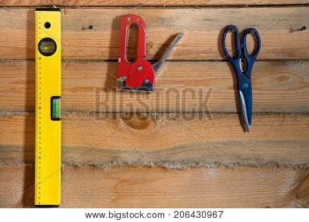Tools for building on wooden background with pine forest. Bright yellow levelred scissors and a stapler hanging on the wall on the screws. Between the pine rail laid with linen tape.