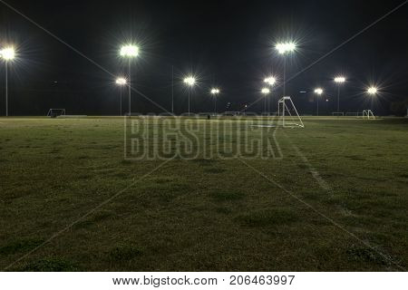 Goal and empty soccer fields at night with the lights illuminating the green grass and reflecting off water drops from a recent rain.