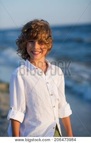 Portrait of boy of 10-11 years against the background of the rough sea. Sea wind ruffled the blond curly hair of the boy. Laddie has blue cheerful eyes suntanned skin a pleasant smile. Boy is dressed in a white shirt.