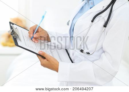 Close-up of a female doctor filling  up medical form at clipboard while standing straight in hospital.