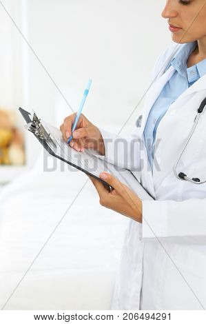 Close-up of a female doctor filling  up medical form at clipboard while standing straight in hospital.