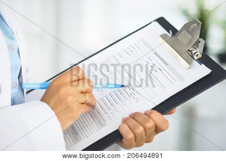 Close-up of a female doctor filling  up medical form at clipboard while standing straight in hospital.