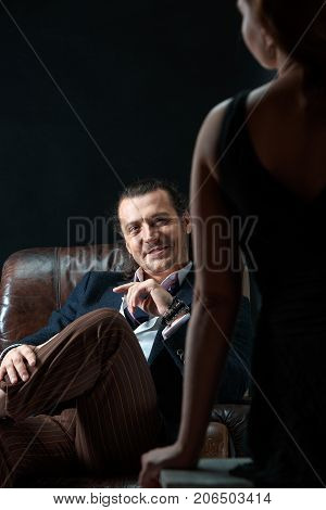 The actor or mature man wearing suit in a classical retro fashion style. The stylish man sitting on a leather couch in a studio. The woman standing near