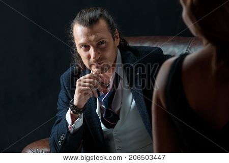 The actor or mature man wearing suit in a classical retro fashion style. The stylish man sitting on a leather couch in a studio. The woman standing near