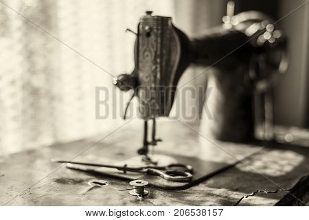 Metal coil with threads in which the needle is inserted and the scissors lie on an old sewing machine against the background of a window through which the sun's rays pass.