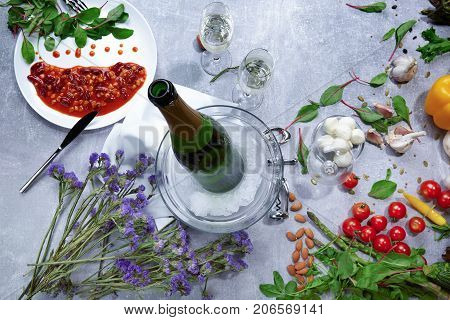 Close-up of a table with a white plate with canned beans with red sauce, a green bottle of champagne, little violet flowers, cloves of garlic, salad pepper, salad leaves on a light gray background.