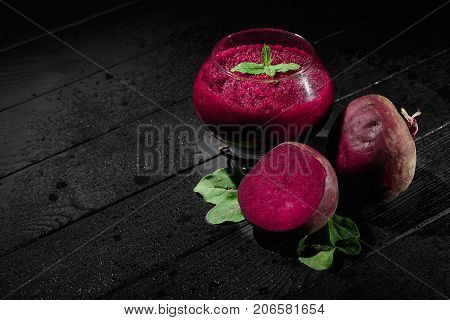 A big, fancy glass filled with saturated red beetroot juice on a black wooden background. Ripe and peeled beets for a healthful summer salad. Raw vegetable cocktails. Copy space.