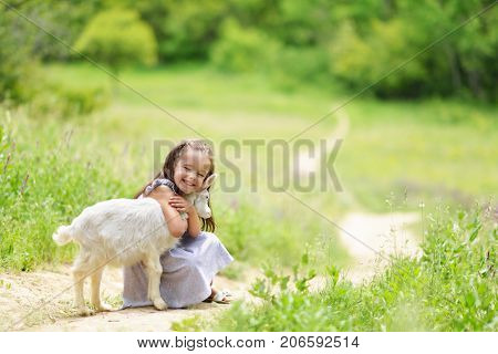 Little girl plays and huhs goatling in country, spring or summer nature outdoor. Cute kid with baby animal, forest, trod, glade background. Friendship of child and yeanling, image toned.