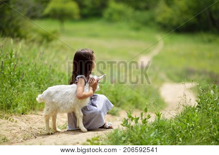 Little girl plays and hugs goatling in country, summer nature outdoor. Cute kid with baby animal, countryside outdoor portrait, forest, trod, glade background. Friendship of child and yeanling