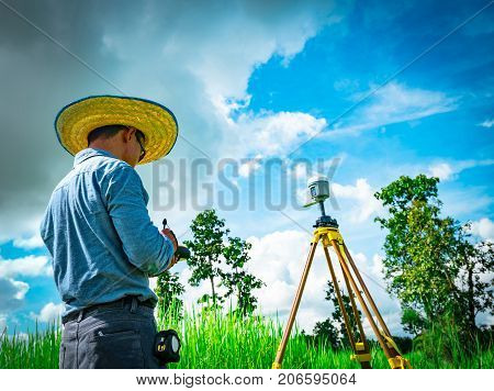 Asian smart engineer or surveyor in black jeans and long sleeve shirt and woven bamboo hat. He is working on controller screen for surveying land in rice field Thailand. GPS surveying instrument.