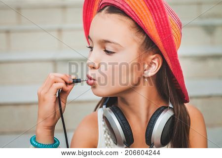 funny girl puts the plug into her mouth. child in a red hat, white blouse and with grey headphones on the background of stairs