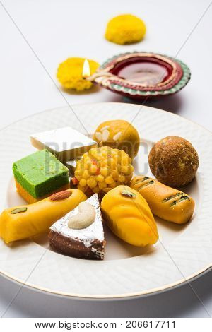 stock photo of Indian sweet or mithai and oil lamp or diya with gift box and flowers on decorative or colourful background, selective focus