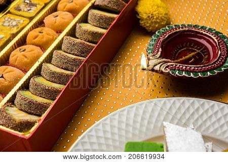 stock photo of Indian sweet or mithai and oil lamp or diya with gift box and flowers on decorative or colourful background, selective focus