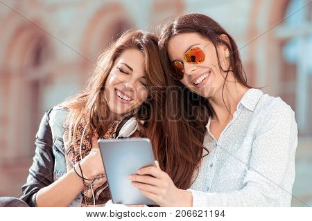 Portrait of two beautiful young woman sitting on bench looking at tablet and have fun.