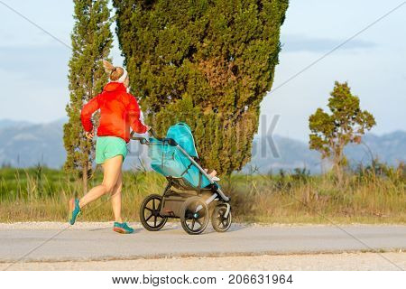 Running mother with child in stroller enjoying motherhood at sunset and mountains landscape. Jogging or power walking woman with pram on at sunset. Beautiful inspirational mountains landscape.