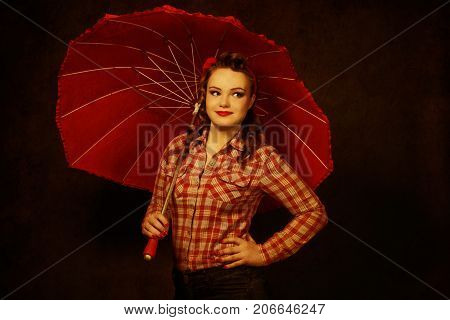 Young pretty pinup girl red button shirt looking aside dark color background retro vintage 50's style with red umbrella.