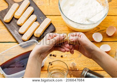 Picture of tiramisu cooking, human hands with savoyardi cookies, coffee, cups, cheese on wooden table