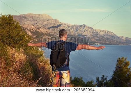 Backside view of grey haired stylish mature man traveler on hill with backpack spreading his arms feeling free happy and relaxed enjoying nice summer day while spending weekend abroad at seaside.