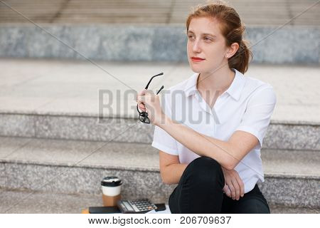 Portrait of thoughtful young Caucasian businesswoman holding glasses sitting on staircase. Work life balance concept
