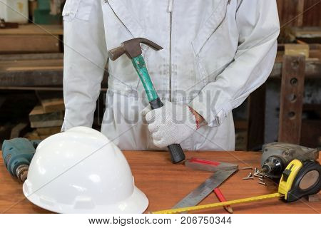 Professional worker with safety uniform holding hammer with other tools on wood workbench in carpentry workshop.