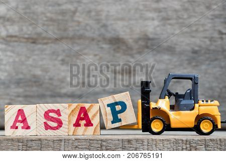 Yellow plastic forklift hold letter P to complete word ASAP (abbreviation of as soon as possible) on wood background